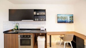 Una cocina compacta con horno integrado, un lavamanos y un pequeño cubo de basura. Hay una mesa de comedor de madera con una silla y un televisor de pantalla plana montado en la pared.