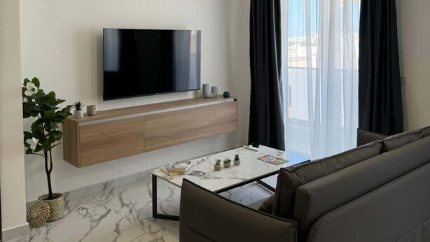 A modern living room with a flat-screen TV mounted on a wooden console, a marble-tiled floor, a leather sofa, and a dining table with a few items on it. - Modern 1-Bedroom Apartment with Balcony – by HostMT (San Pawl il-Baħar)