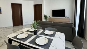 A modern living room with a marble-topped dining table, grey chairs, a grey sofa, a flat-screen TV, and a wooden cabinet. - Modern 1-Bedroom Apartment with Balcony – by HostMT (San Pawl il-Baħar)