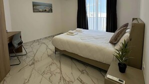A hotel room with a bed, a desk, a chair, a potted plant, and a painting on the wall. - Modern 1-Bedroom Apartment with Balcony – by HostMT (San Pawl il-Baħar)