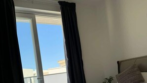A hotel room with a large bed, two rolled towels, a small potted plant, and a view of the sky through a window with black curtains. - Modern 1-Bedroom Apartment with Balcony – by HostMT (San Pawl il-Baħar)