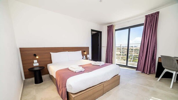 Habitación de hotel con una cama grande, una mesita de noche pequeña, una silla y vistas de la ciudad a través de un ventanal amplio.