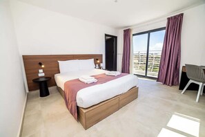 Habitació d'hotel amb un llit gran, una taula de nit petita, una cadira i vista a la ciutat a través d'una finestra ampla.