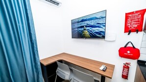 Une pièce avec un bureau en bois, un téléviseur fixé au mur qui diffuse une image d’un kayak sur l’eau, une couverture anti-feu, une trousse de premiers secours et un extincteur accrochés au mur.