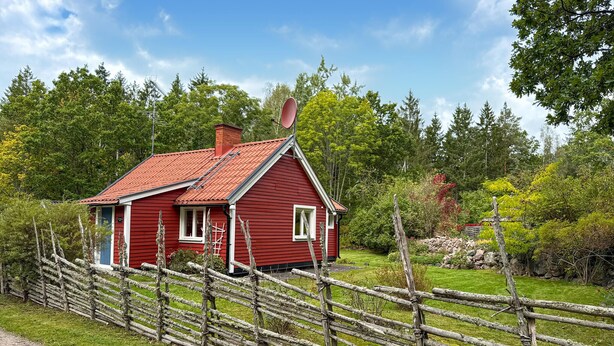 Une maison en bois rouge, avec un toit en pente, entourée d’une clôture en bois et d’une végétation luxuriante.