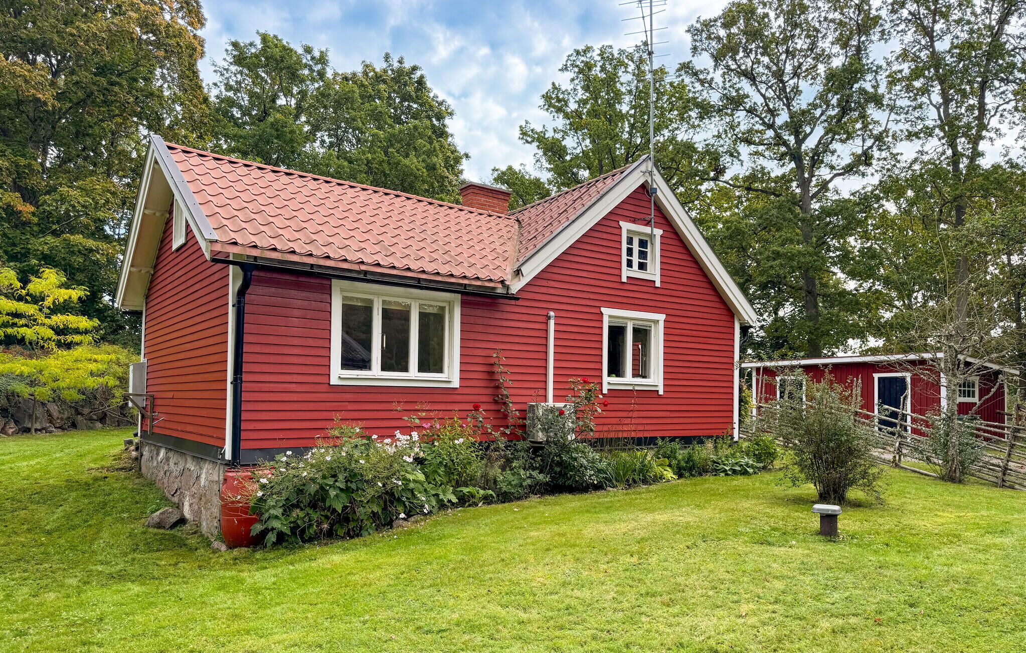A red wooden house with a sloped roof, white window frames, and a small garden with various plants and a bench.