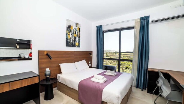 Quarto de hotel com cama, escrivaninha, cadeira, mesa pequena e janela com cortinas.