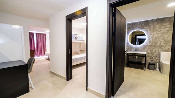Ein Hotelzimmer mit Bett, Schreibtisch, Stuhl, einem Spiegel mit rundem Rahmen sowie einem Badezimmer mit Waschbecken und Toilette.