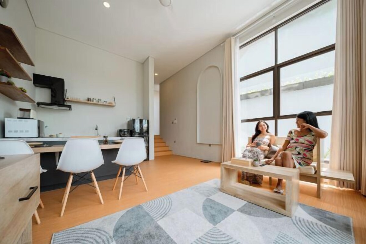 A modern living room with a large window, a kitchen area, and a seating area with two people.