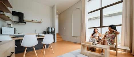 A modern living room with a large window, a kitchen area, and a seating area with two people.