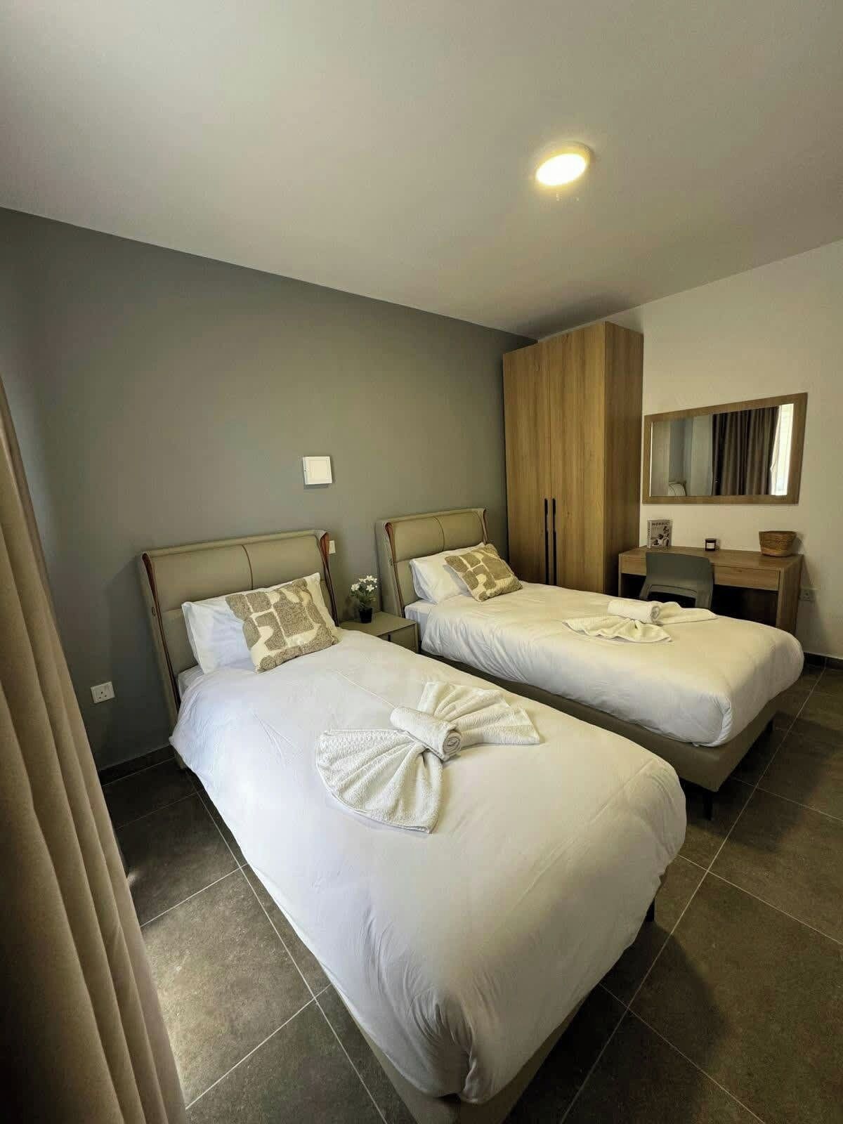 A hotel room with two beds, a wooden wardrobe, a desk with a chair, and a mirror on the wall.