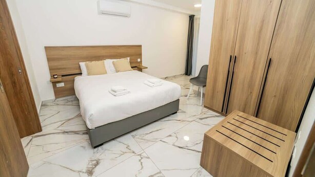A modern hotel room with a marble floor, a wooden bed frame, a wooden wardrobe, and a small wooden bench. - Luxury 3-Bedroom Family Maisonette – by HostMT (San Pawl il-Baħar)