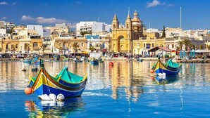 Un pueblo costero con coloridas embarcaciones amarradas en el agua, una iglesia destacada con un campanario y un cielo azul despejado.