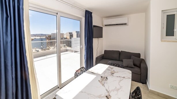 A modern room with a large sliding glass door leading to a balcony, a grey sofa, a marble table, and an air conditioning unit on the wall. - Bright Penthouse with Private Terrace by HostMT (San Pawl il-Baħar)