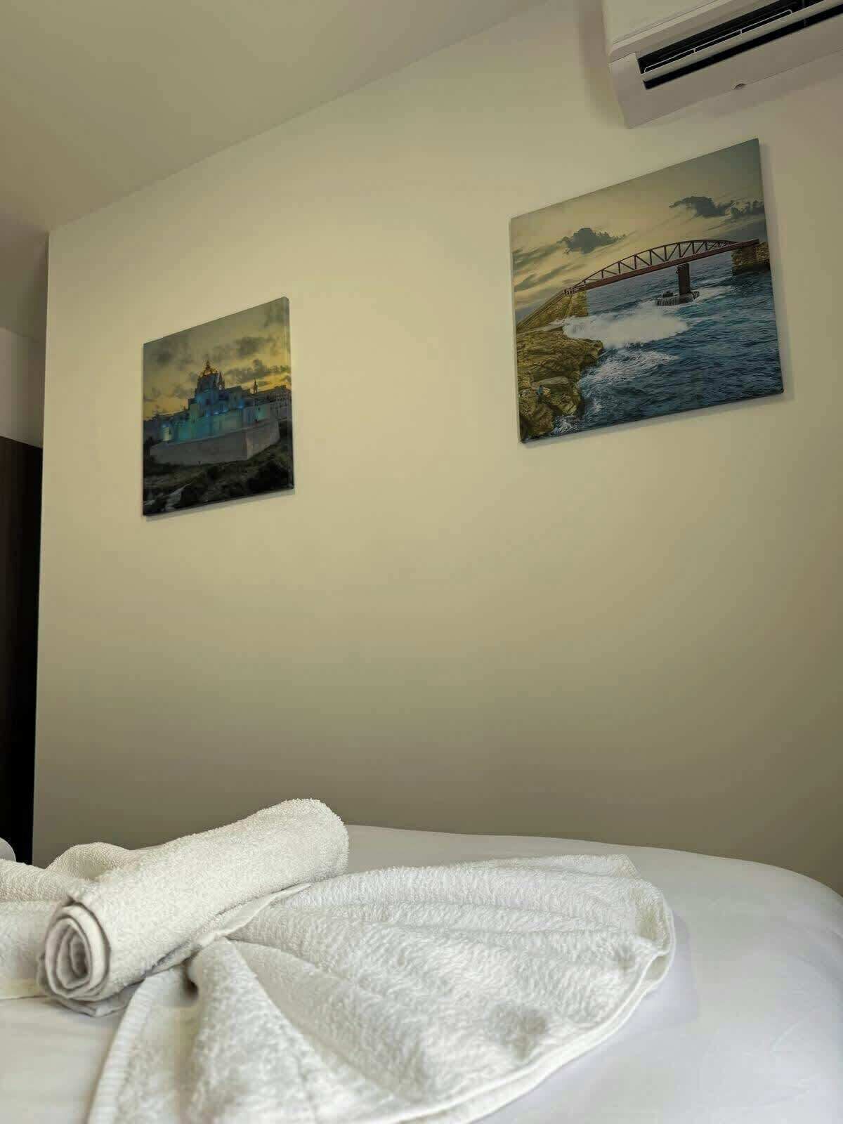 A hotel room with a bed, a rolled white towel, and two paintings on the wall depicting a bridge and a building.