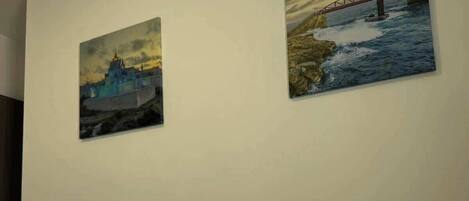 A hotel room with a bed, a rolled white towel, and two paintings on the wall depicting a bridge and a building.