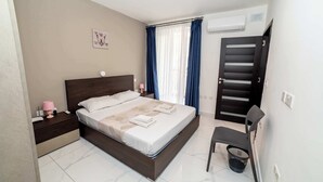Un dormitorio ordenado con cama, mesitas de noche, una silla y una ventana con cortinas.