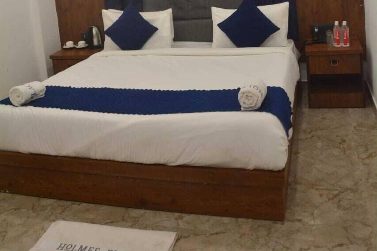A hotel room with a wooden bed frame, a headboard with a grey and blue panel, white bedding, and a sign on the floor reading "HOLMES PLACE".