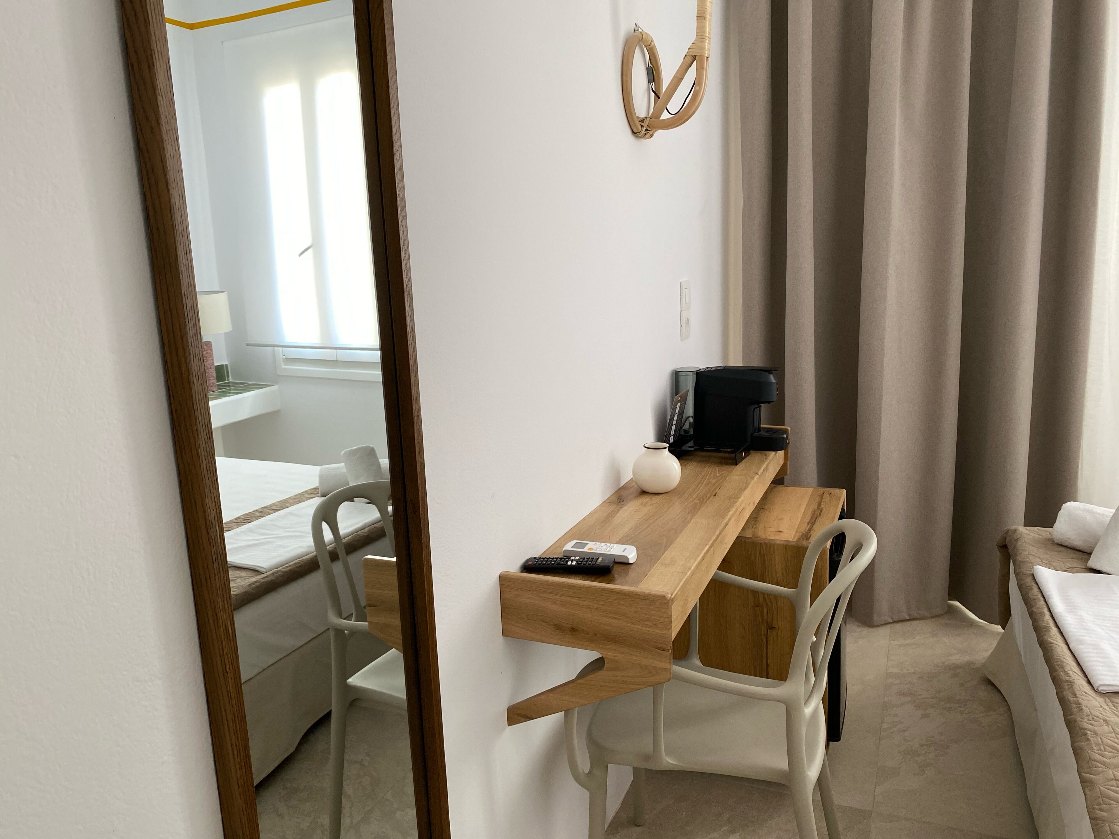 A hotel room with a wooden desk, white chairs, a bed with beige bedding, a coffee machine, and a mirror.
