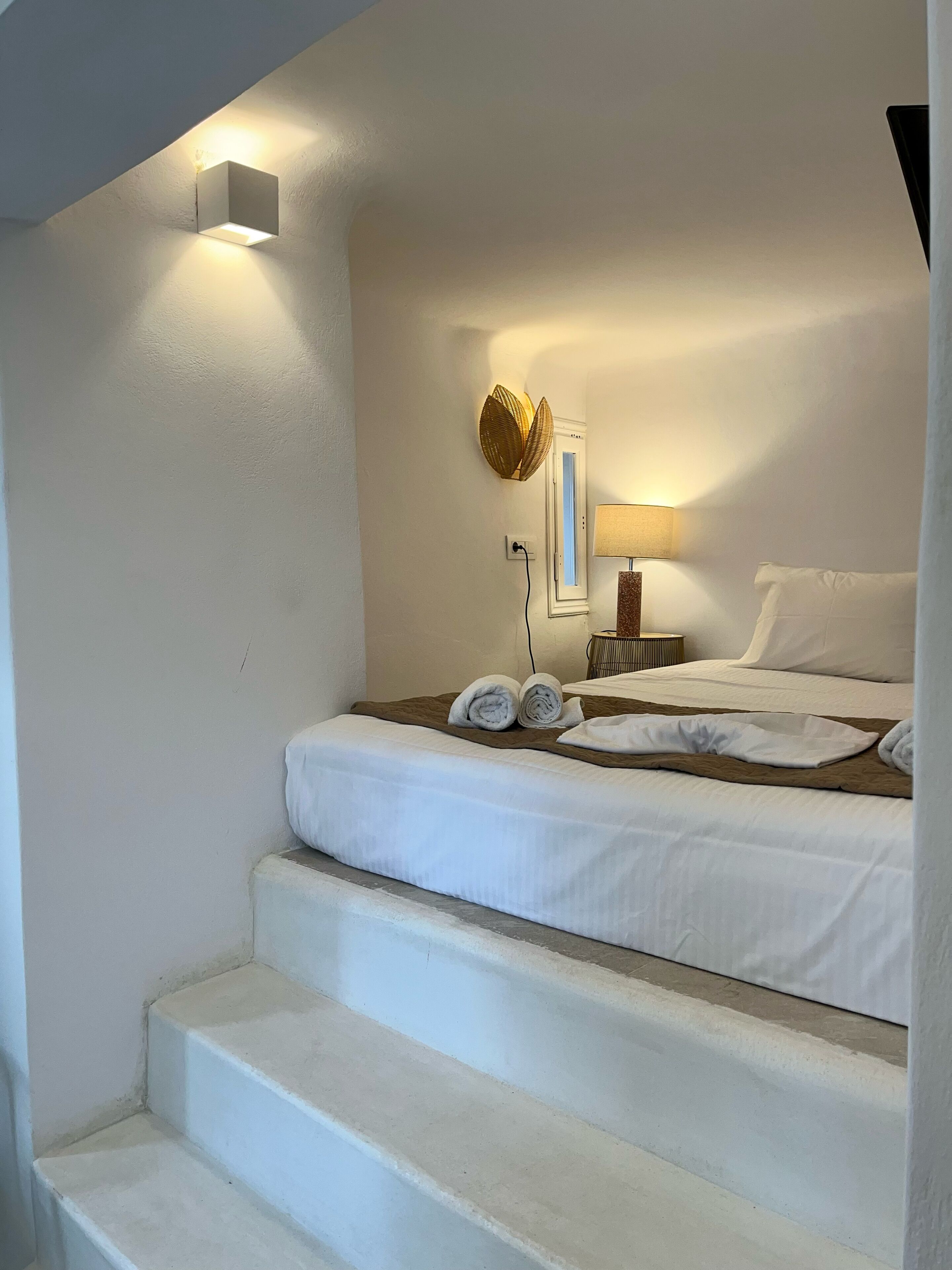 A bedroom with a bed, a nightstand, a lamp, a wall-mounted light, and a window.