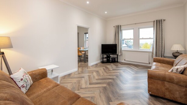 Een moderne woonkamer met een visgraat houten vloer, twee bruine leren banken, een flatscreen televisie en een eethoek op de achtergrond.