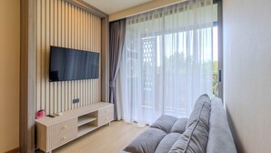 Een moderne woonkamer met een flatscreen-tv aan een houten paneelwand, een grijze bank en een groot raam met dunne gordijnen.