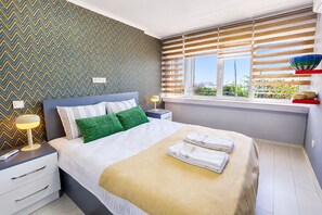 A modern bedroom with a bed, nightstands, a lamp, a window with blinds, and a view of greenery outside.