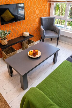Un salon moderne avec un canapé gris, une table basse ornée de bougies, un téléviseur fixé au mur et un mur orange à motifs.
