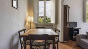 Een eethoek met een houten tafel en stoelen, een raam met geopende luiken, een lamp, een televisie en een kast.