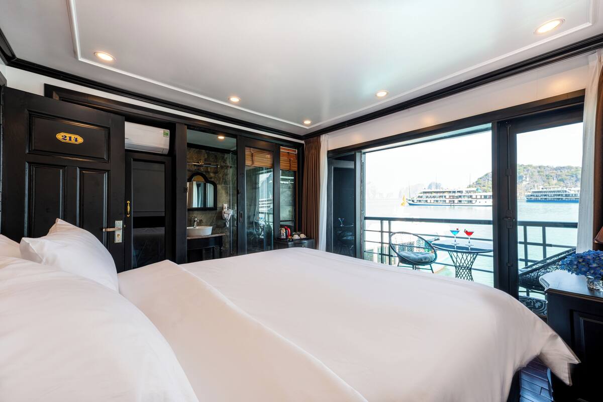 A hotel room with a large bed, a bathroom with a glass door, and a balcony with a view of a body of water and a boat.