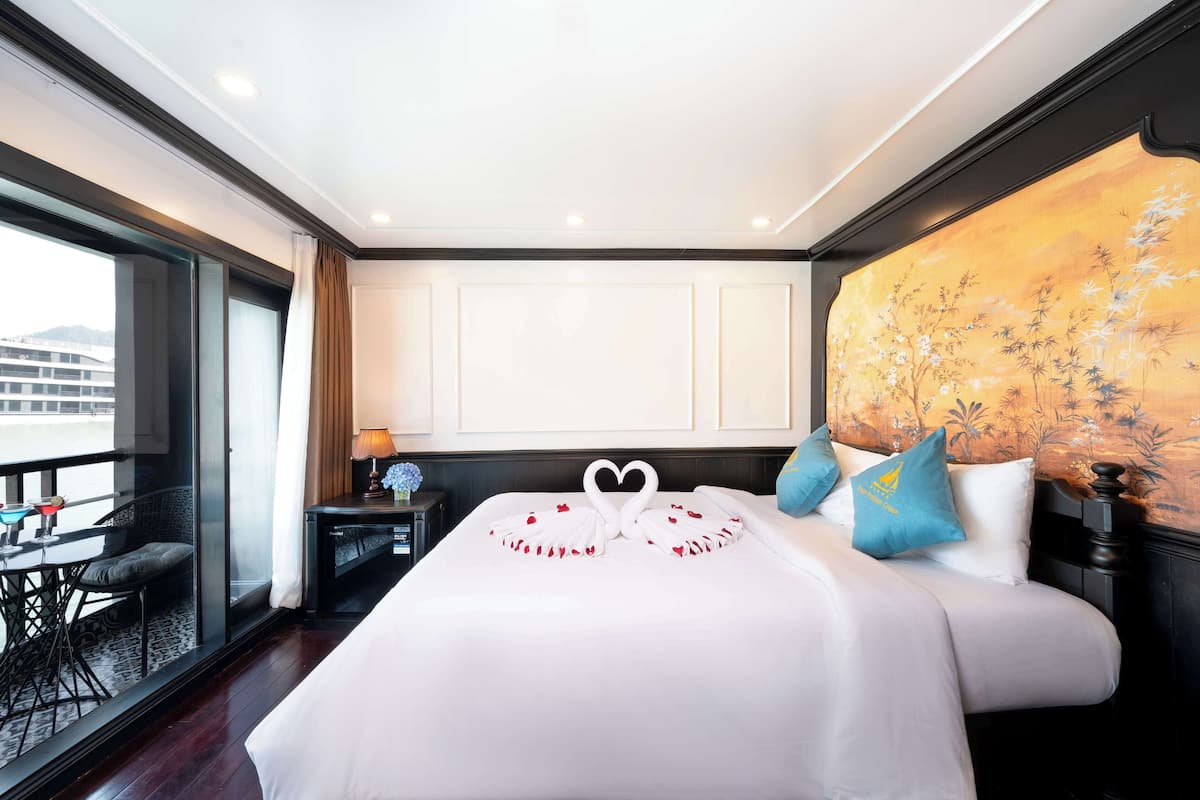 A hotel room with a large bed, a balcony with a table and chairs, and a painting on the wall.