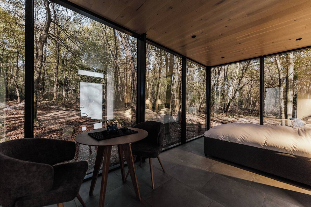 A modern bedroom with a large bed, a dining table, two chairs, and a view of a forest through large windows.