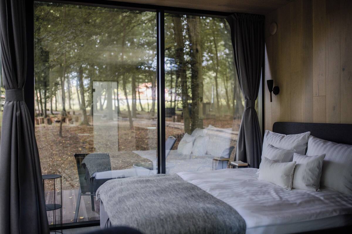 A bedroom with a large window, a bed with multiple pillows, a chair, and a small table.