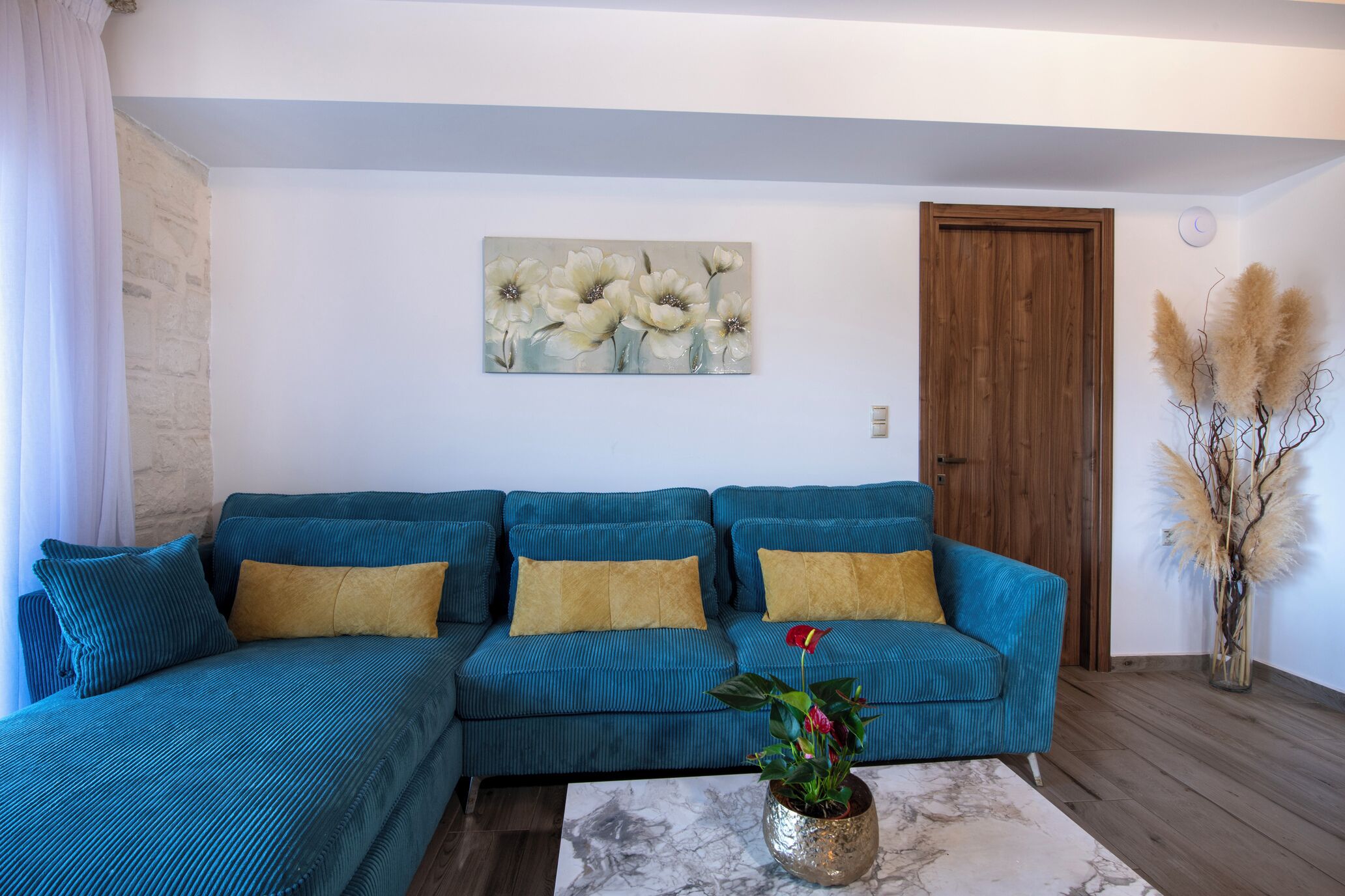 A teal sofa with yellow cushions, a marble coffee table, a painting of flowers, and pampas grass in a vase.