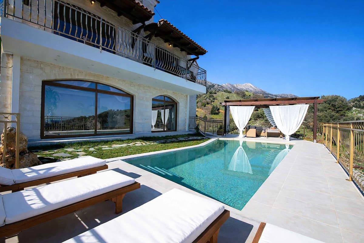 Ein Poolbereich mit Liegestühlen, einer Pergola mit weißen Vorhängen und einem Blick auf die Berge im Hintergrund.
