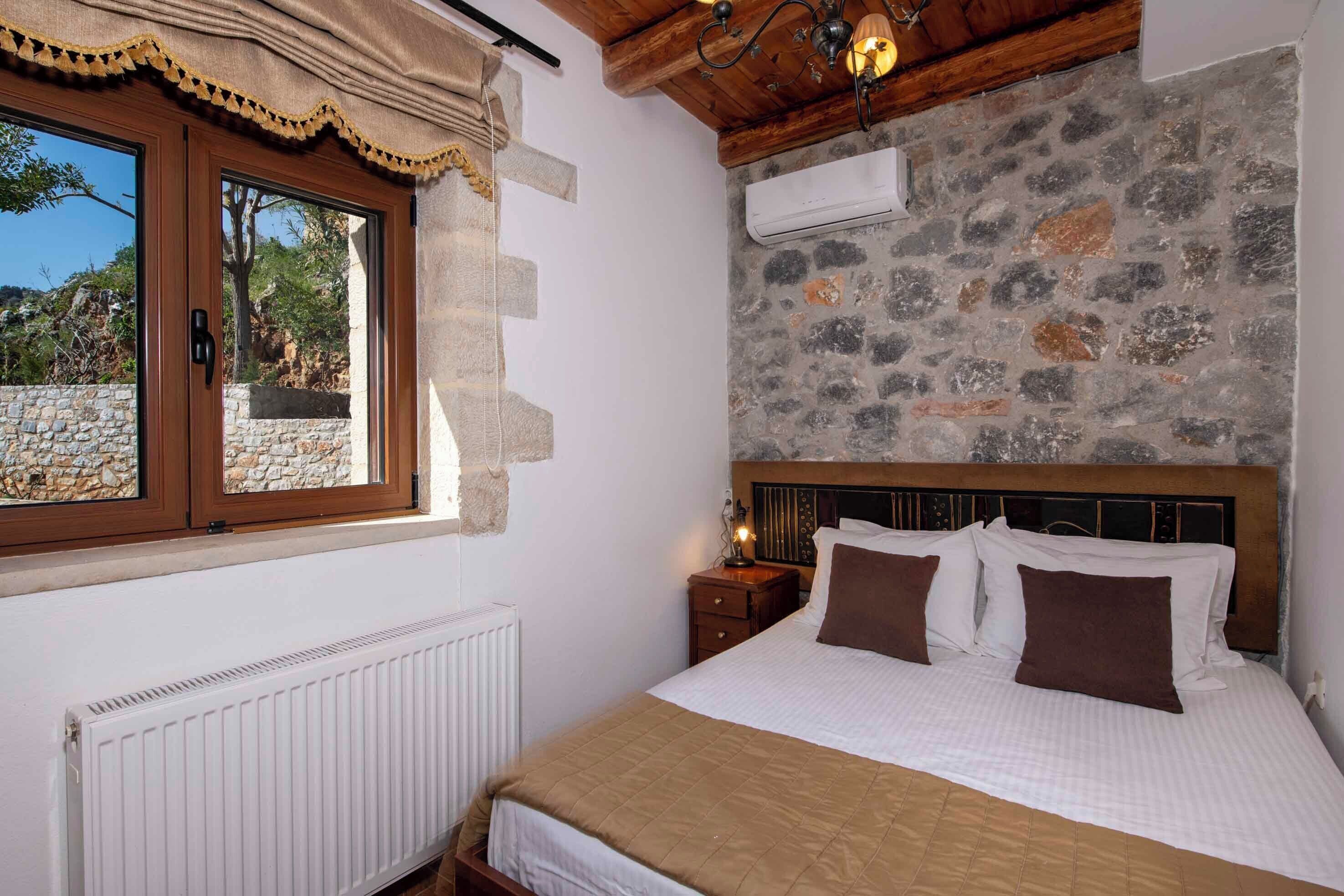 A bedroom with a stone wall, a wooden ceiling, a bed with white and brown bedding, and a window with curtains.