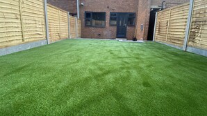 A backyard with artificial grass, a brick house, and wooden fences.