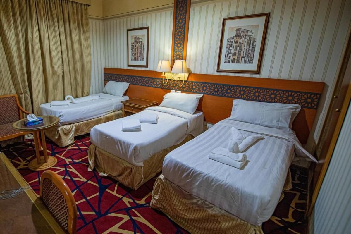 Habitación de hotel con tres camas individuales, una mesita pequeña con una silla y una alfombra estampada.