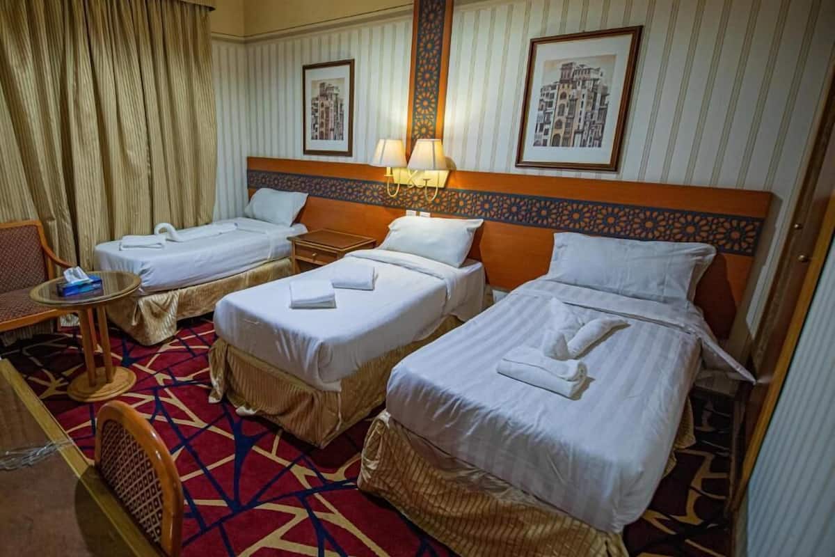 Habitación de hotel con tres camas individuales, una mesita pequeña con una silla y una alfombra estampada.