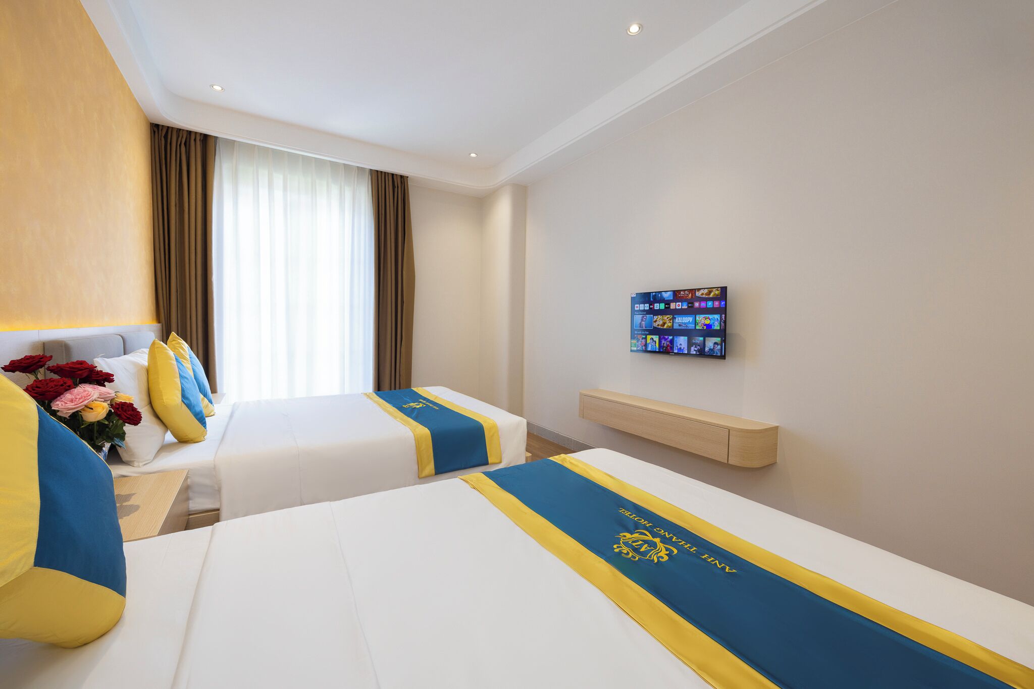 Habitación de hotel con dos camas, cada una con cubrecama azul y amarillo, cabecero de madera, mesita de noche con un florero y televisor montado, además de un estante de madera.