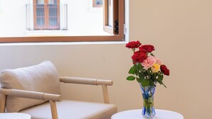 Una habitación con ventana, una silla, una mesita y un florero con flores.