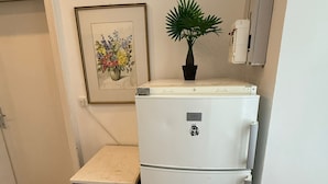 Uma cozinha compacta com geladeira branca, um armário pequeno de cor escura, uma obra de arte floral emoldurada e uma planta em vaso na bancada.