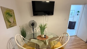Uma sala de estar moderna com uma mesa de vidro redonda, cadeiras de vime branco, uma televisão de tela plana na parede e um pequeno ventilador.