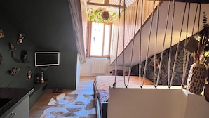 A narrow hallway with a staircase leading to a second floor. The hallway features a patterned rug, a bed with a headboard, and a hanging plant.