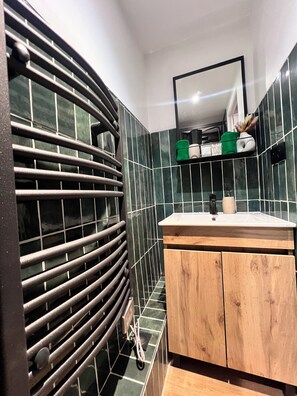 A bathroom with a black radiator, green tiled walls, a wooden vanity with a mirror, and a towel rack.