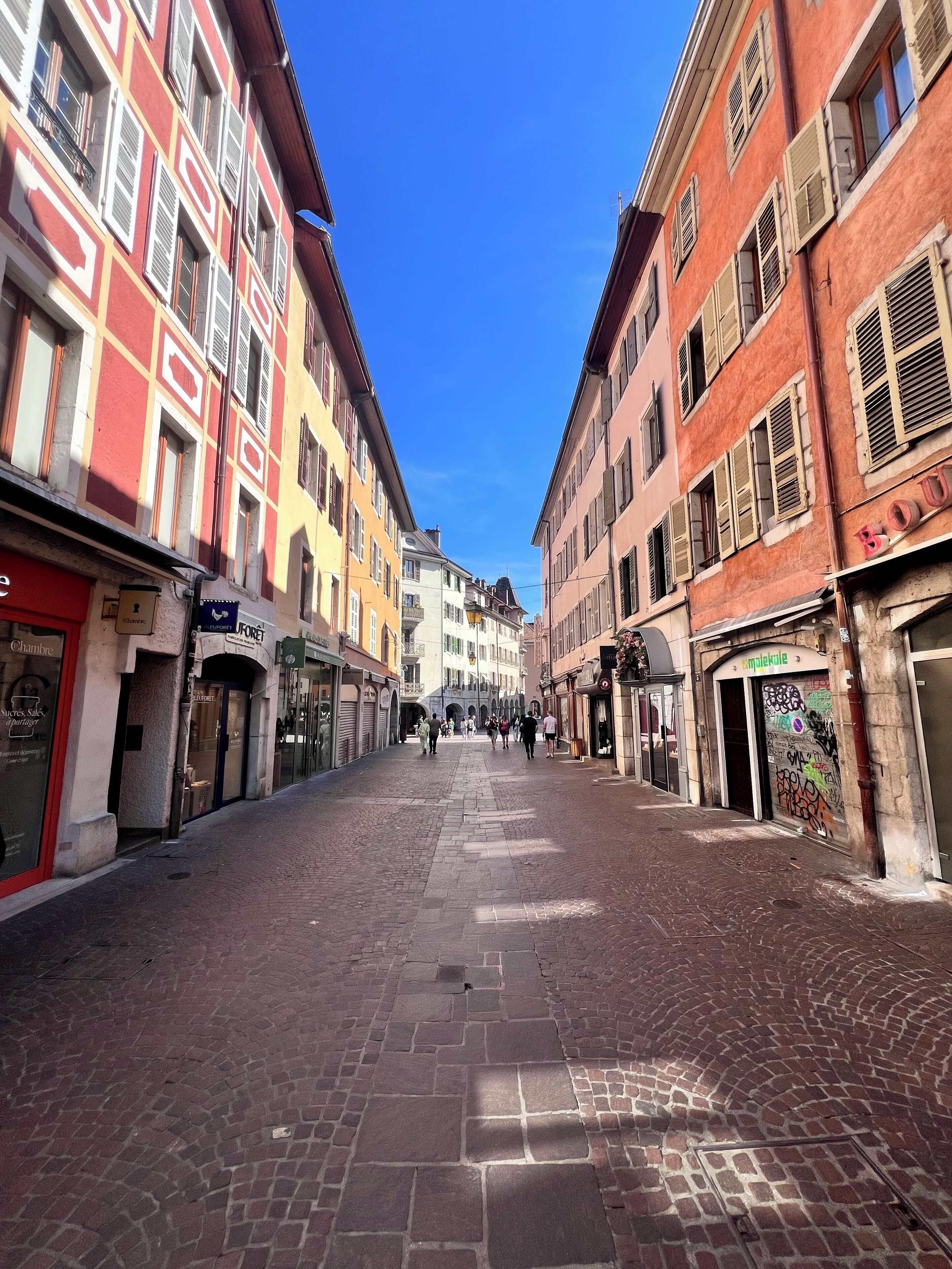 Une rue pavée bordée d’immeubles à plusieurs étages, chacun abritant différents commerces, dont une boutique dont l’enseigne indique clairement « Chambre ».
