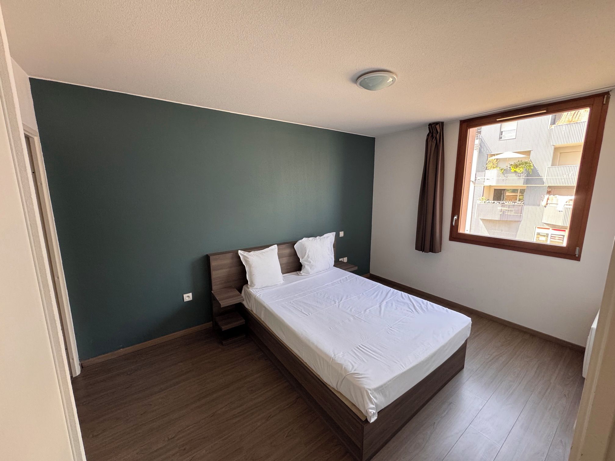 Une chambre à coucher avec un lit, une table de chevet, une fenêtre avec des rideaux et une vue sur les bâtiments à l’extérieur.