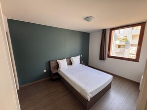 Een slaapkamer met een bed, een nachtkastje, een raam met gordijnen en uitzicht op gebouwen.