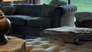 Un salon chaleureux avec un canapé gris, une table d’appoint en bois et une grande fenêtre donnant sur un champ verdoyant et un ciel nuageux.
