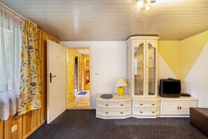 Et værelse med en trædør, et hvidt skab med skuffer, et fjernsyn på et stativ og en lampe på et lille bord.